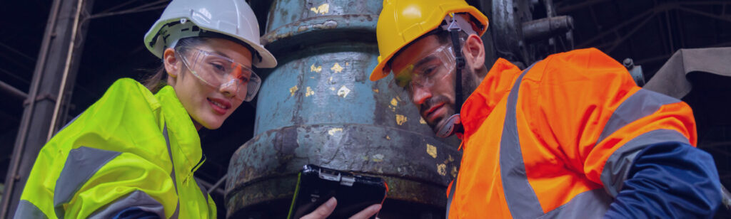 Dois profissionais da indústria com EPIs e capacetes analisando dados em um tablet no chão de fábrica, representando equipes utilizando SCRUM na operação.